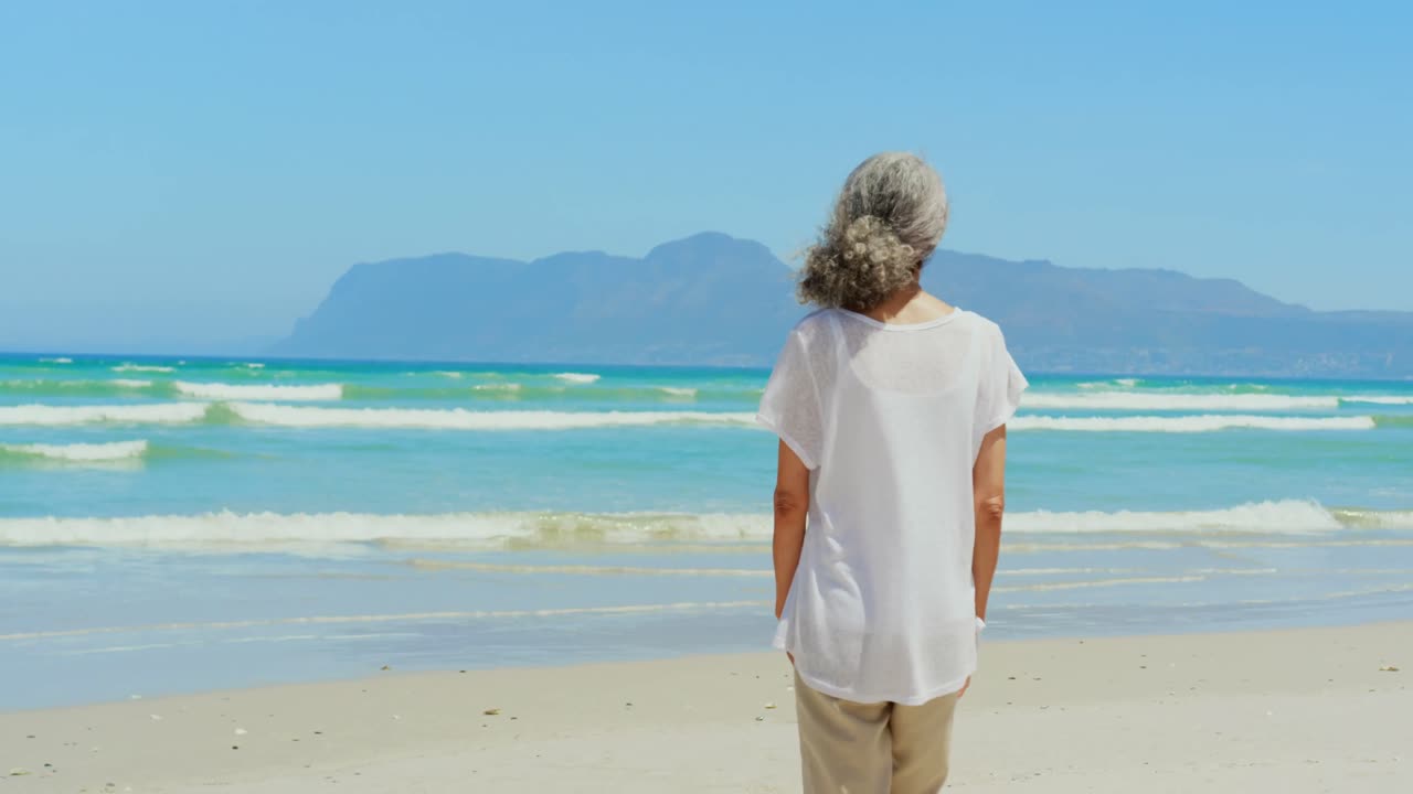vista trasera de una mujer afroamericana senior activa caminando por la playa al sol 4k