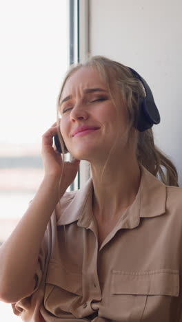 young blonde woman in headphones enjoys popular dynamic music sitting at windowsill leaned on pillow in comfortable room at home slow motion