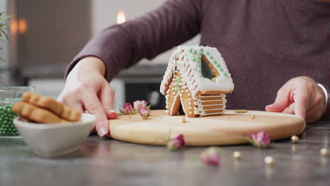 baker carefully rotating and displaying beautifully decorated gingerbread house with white icing and green beads, wooden tray, golden sprinkles, dried flowers, and blurred candlelight in background