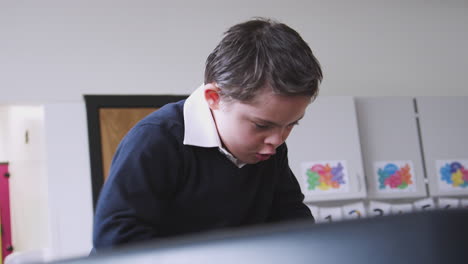 Primary-school-schoolboy-with-Down-Syndrome-standing-at-desk-in-a-classroom-playing,-low-angle