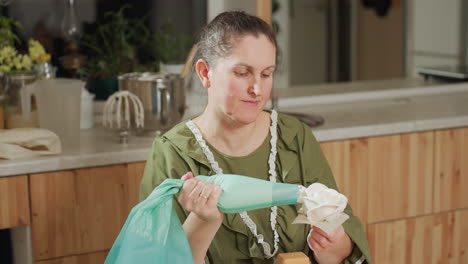 chef in green dress skillfully decorates dessert with white icing in flower shape using piping bag, then sets bag down on table while working in cozy kitchen surrounded by tools and wooden cabinetry