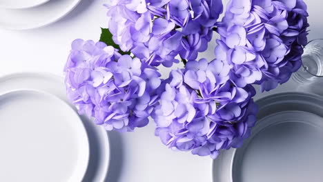 overhead view of purple hydrangeas with white plates and glass on a white background