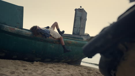 carefree woman lying old boat on beach. girl relax waving foot in black shoe.