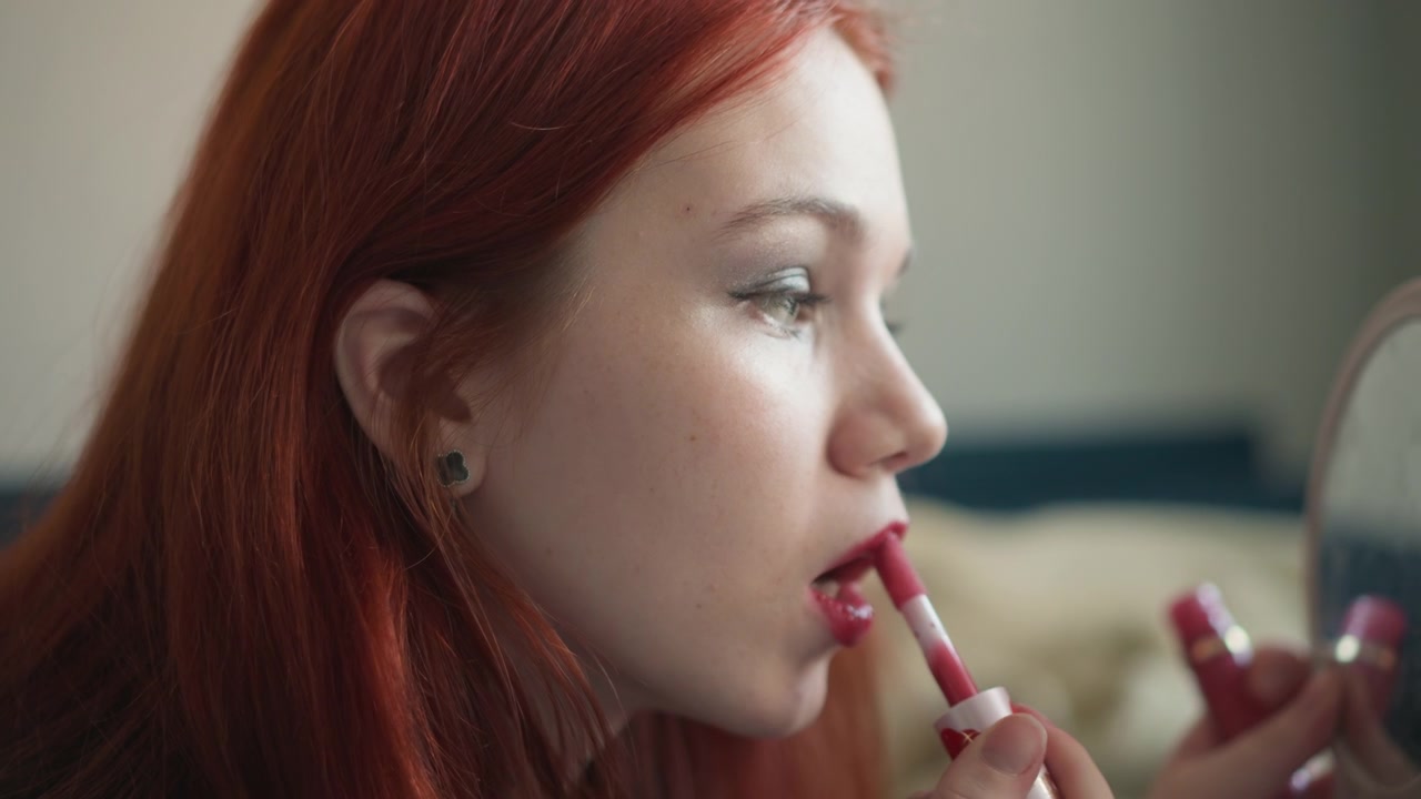Profile of woman with matching lipstick and mirror. Gentle evening reflection of woman with lipstick in window light. Softly illuminated woman with red hair focusing on lipstick reflection by window