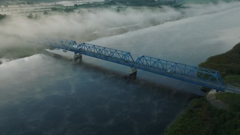 Aerial-establishing-view-of-the-steel-bridge-over-Lielupe-river-on-a-sunny-summer-morning,-fog-rising-over-the-river,-cars-driving,-wide-orbiting-drone-shot
