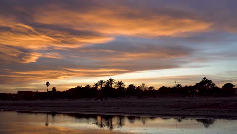 Lapso-De-Tiempo,-Puesta-De-Sol-Con-Nubes-Difusas-Ondeando-Sobre-El-Cielo-Mientras-Dos-Personas-Caminan-Por-La-Arena,-El-Cielo-Muestra-Hermosos-Colores-Dorados-Y-Azules