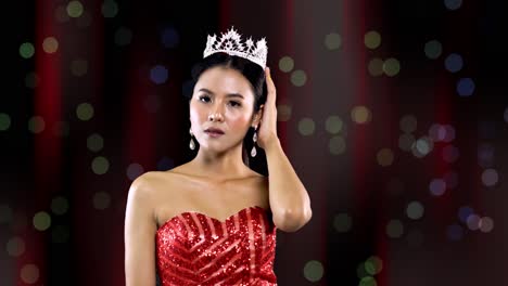 miss beauty pageant queen with asian features and red gown smiles and poses while wearing her crown - stage curtain background with bokeh