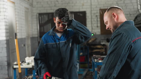 mechanic in blue uniform working on engine while colleague steps back, wipes forehead, then resumes scrapping in busy mechanical shop, workshop filled with tools