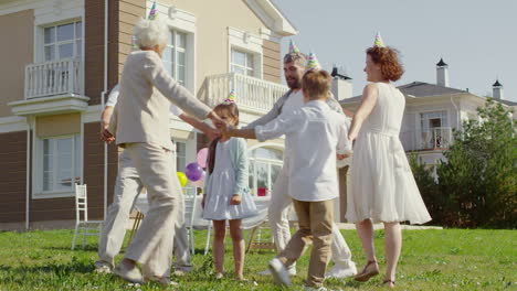 feliz familia multigeneracional con sombreros de fiesta tomándose de la mano y bailando alrededor de una niñita mientras celebraba al aire libre en el patio trasero el día soleado