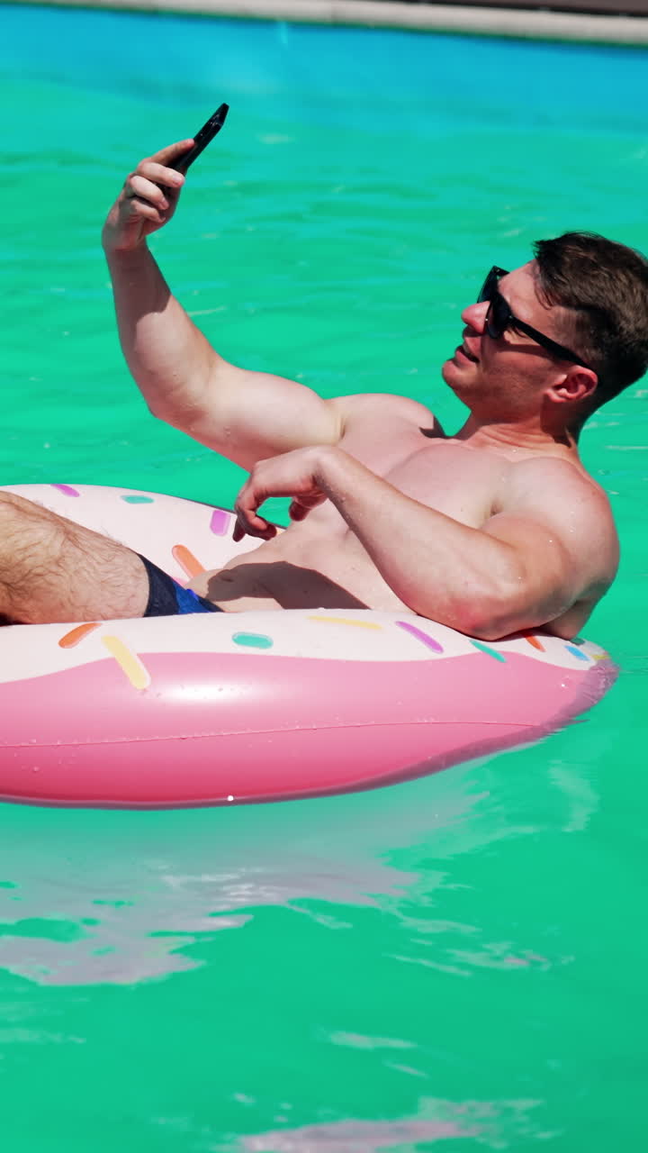 Relaxed Caucasian sportive man swimming in the pool. Man sits in the inflatable donut having a video chat on his phone. Vertical video