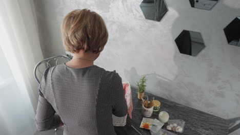 aerial view of lady in checkered dress retrieving item from shopping basket placing it on table with wall mirror reflecting her and another person, nearby chair and visible wall outlet in kitchen