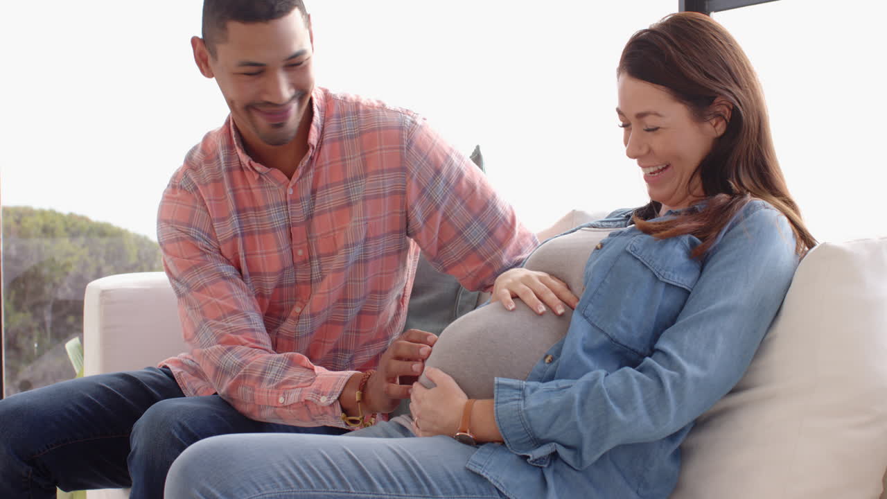 Expecting couple sitting on couch, man touching pregnant woman's belly lovingly