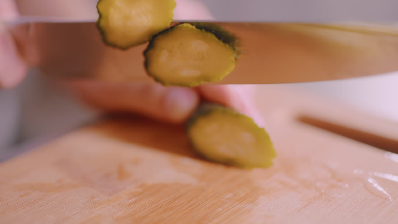 Close up of hand slicing cucumber with sharp knife on wooden cutting board, fresh vegetable being prepared in kitchen for meal, showing detail of texture and precision of cutting process
