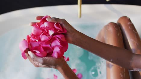 african american woman taking bath and touching flower petals in water in bathroom