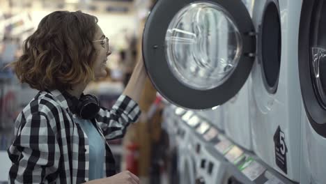 una mujer joven positiva en una camisa a cuadros eligiendo la lavadora en la tienda de electrodomésticos. abre la puerta y mira adentro. vista lateral