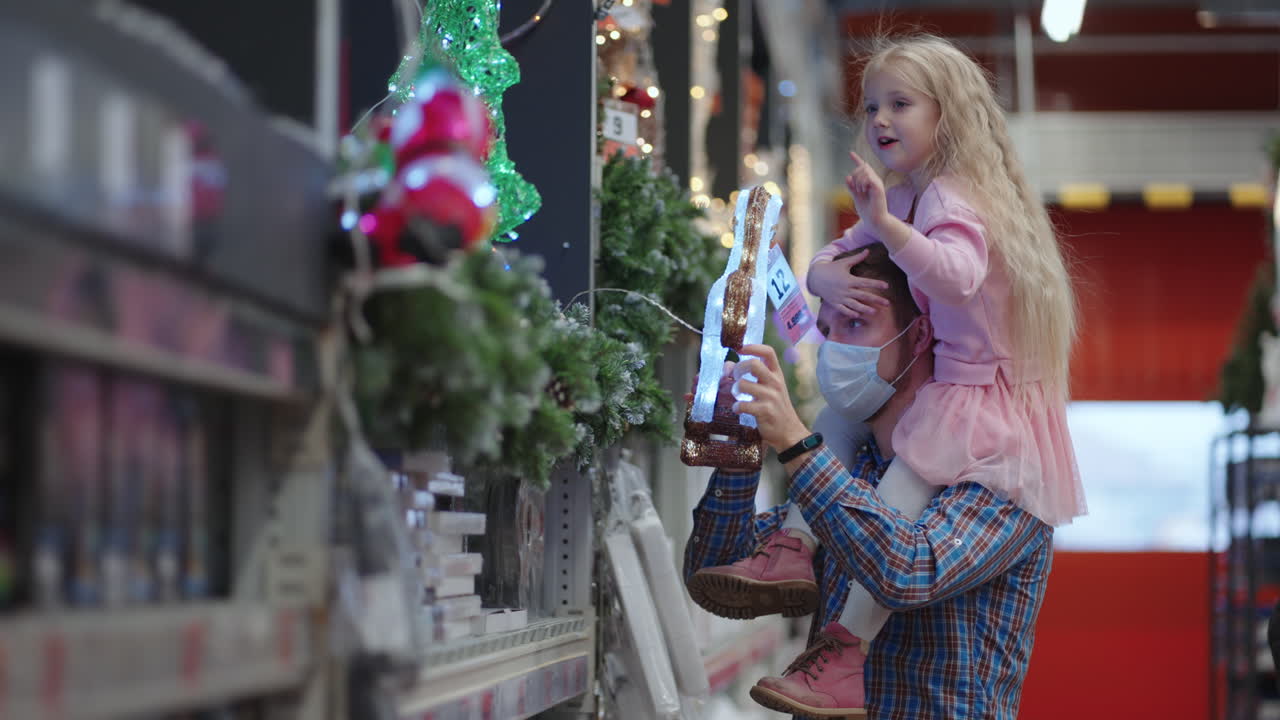 папа и дочка выбирают украшения и гирлянды с игрушками для новогодних елок и дома в магазине. новогодние гирлянды и декор