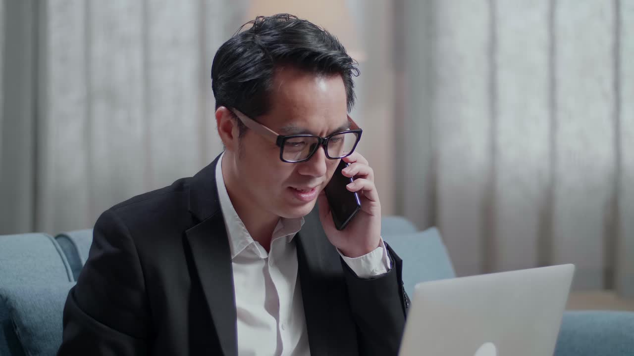재과 쇼트를 입은 아시아 사업가가 집에서 노트북으로 일하면서 전화에 응답하고 전화를 하고 있습니다.