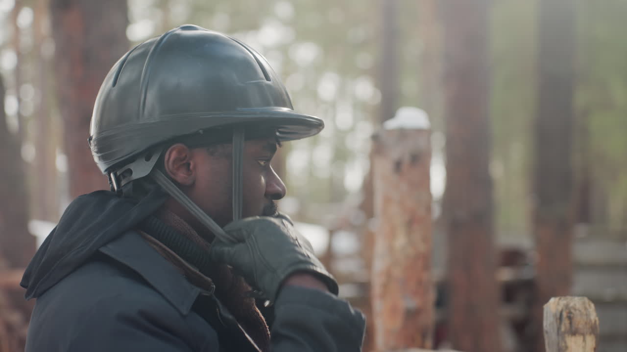 preparación para un ritual ecuestre, persona en establo ajustando el equipo antes de que empiece la sesión de monta, jinete preparando meticulosamente el casco y los guantes antes de comenzar a montar en un establo rodeado de bosques