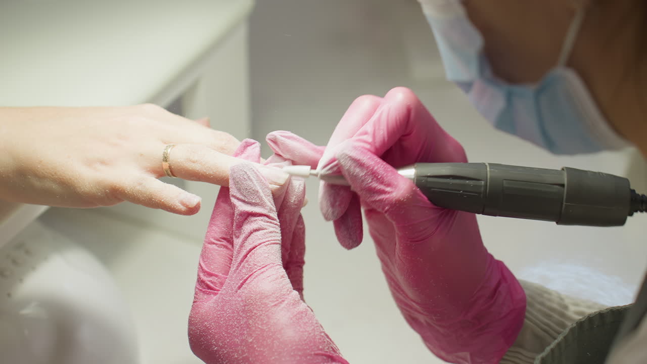 Close-up view of nail technician in pink gloves filing client nail with electric tool an technician's masked face partially visible and client also wears gold ring and rests hand on white surface