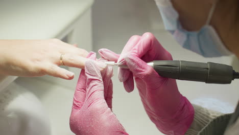 close-up view of nail technician in pink gloves filing client nail with electric tool an technician's masked face partially visible and client also wears gold ring and rests hand on white surface