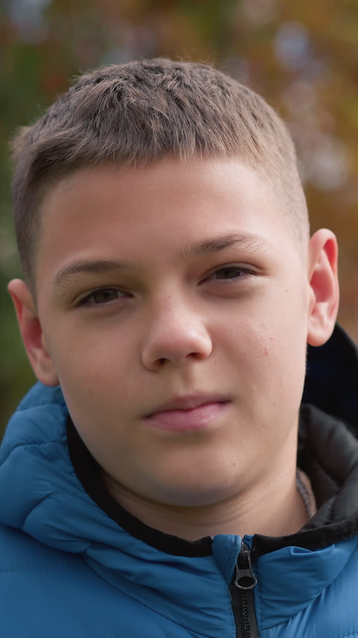niño con chaqueta azul al aire libre con una sonrisa sutil y la cabeza inclinada ligeramente, rodeado de árboles de otoño vibrantes, casa borrosa de techo rojo, y la luz natural suave