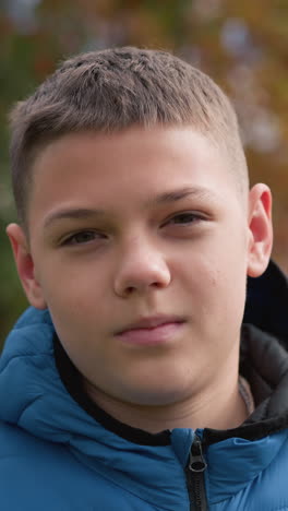 niño con chaqueta azul al aire libre con una sonrisa sutil y la cabeza inclinada ligeramente, rodeado de árboles de otoño vibrantes, casa borrosa de techo rojo, y la luz natural suave