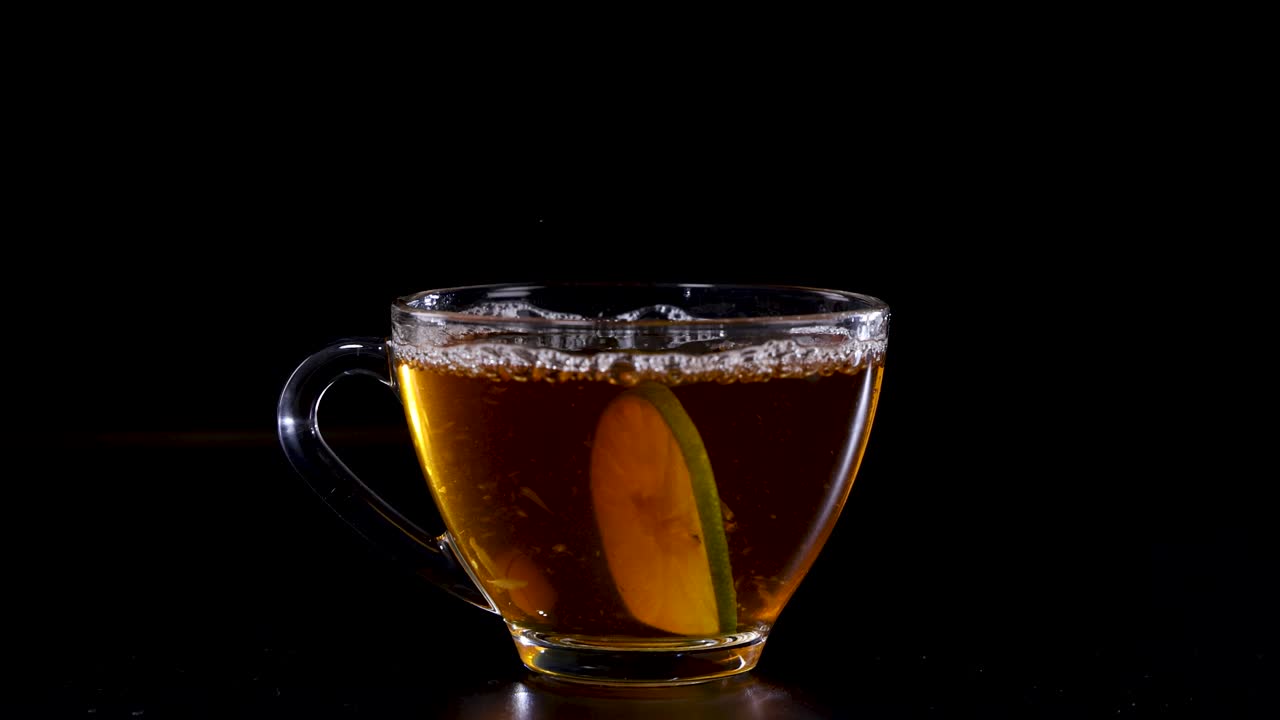 A lime slice drops into a glass cup of tea, creating ripples against a dark backdrop with dramatic lighting