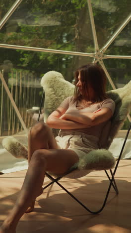 thoughtful woman with crossed hands sits in armchair. lady relaxes at panoramic terrace window. female relishes overlooking forest landscape in calm atmosphere