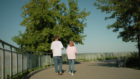 lady in pink top and jeans walking alongside friend holding laptop in left hand near iron railing on paved path surrounded by greenery, under clear sky