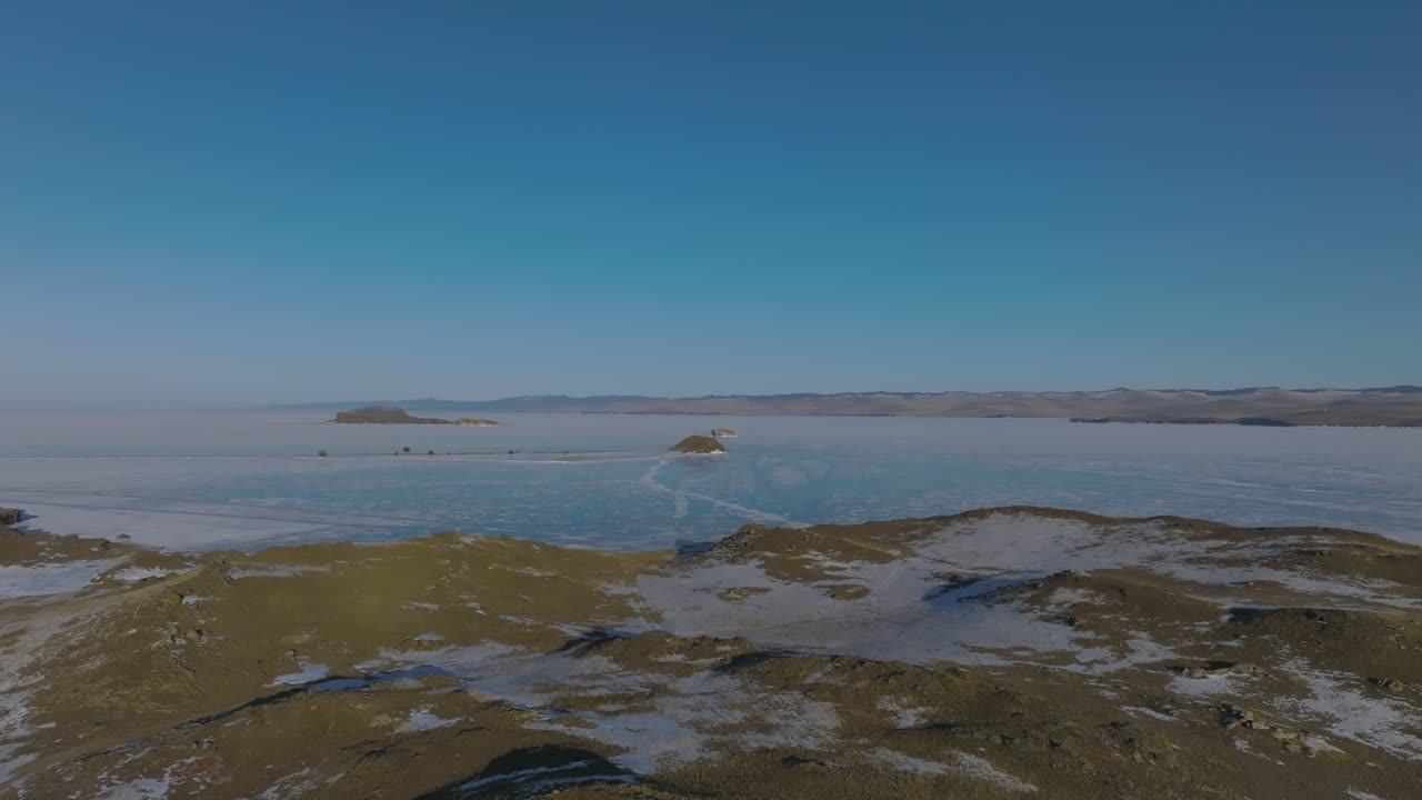 Bredt luftopptak av det frosne Maloye More-stredet på Bajkalsjøen, Irkutsk oblast, Russland, med øyer, snøflekker og klar blå himmel på en rolig vinterdag.