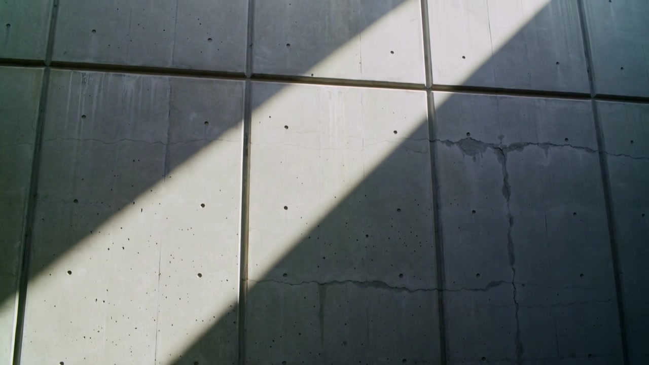 Cámara haciendo un paneo que revela una banda diagonal de luz solar en una pared de paneles en un estacionamiento, mostrando juntas y manchas.