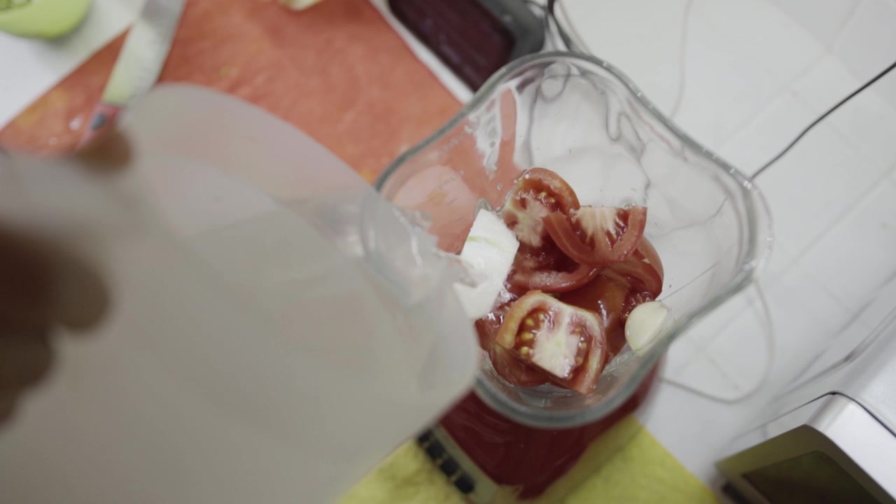 Vista dall'alto dell'acqua versata nel boccale del frullatore con pomodori tritati e ingredienti, preparando il composto per la cottura con consistenze fresche e un semplice processo di cucina.