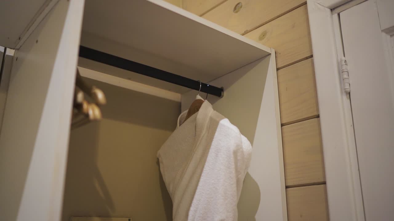 una toma de panorámica lenta revela un armario de túnicas en una habitación de hotel, con una túnica de baño blanca bien colgada, invitando a los huéspedes a relajarse y disfrutar de su estancia en comodidad y lujo