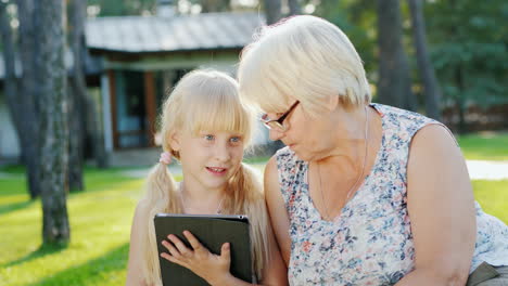 Großmutter-Und-Enkelin-Ruhen-Sich-Im-Hinterhof-Des-Hauses-Aus,-Sie-Schauen-Gemeinsam-Auf-Das-Tablet