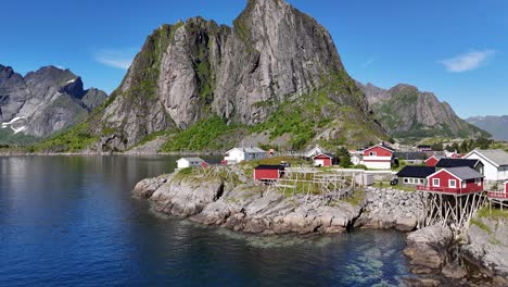 drone flying under the car bridge and over small fishing village hamnøy in norway towards iconic mountains above the sea with wooden drying racks for torrfisk