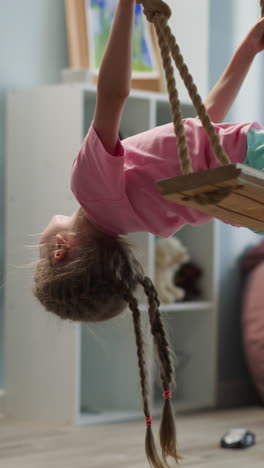 girl with long braids swings holding ropes in cozy apartment. female preschooler has fun at home. child plays in room with bookshelf and pictures