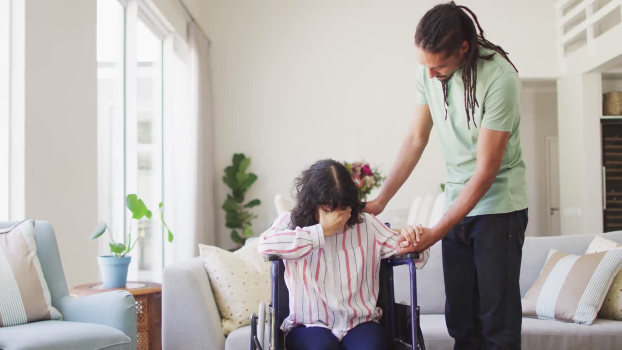 hombre biracial tomándose de la mano y consolando a una mujer triste en silla de ruedas en la sala de estar