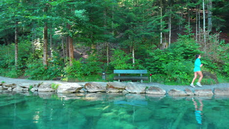 Drone-shot-of-older-sporty-woman-stretching-by-a-beautiful-lake-in-nature