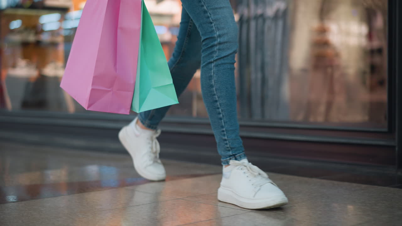 vista de la pierna de una persona con vaqueros y zapatillas de deporte de lona blanca sosteniendo bolsas de compras rosadas y menta mientras camina a través de un centro comercial más allá de una tienda de ropa, con fondo borroso de pantallas minoristas