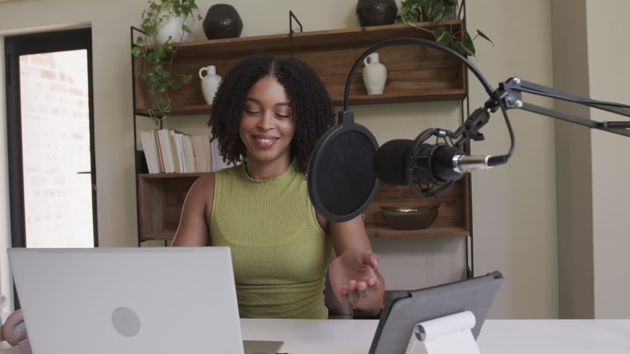 Home studio African American woman recording talking into mic fixing pop shield using laptop tablet