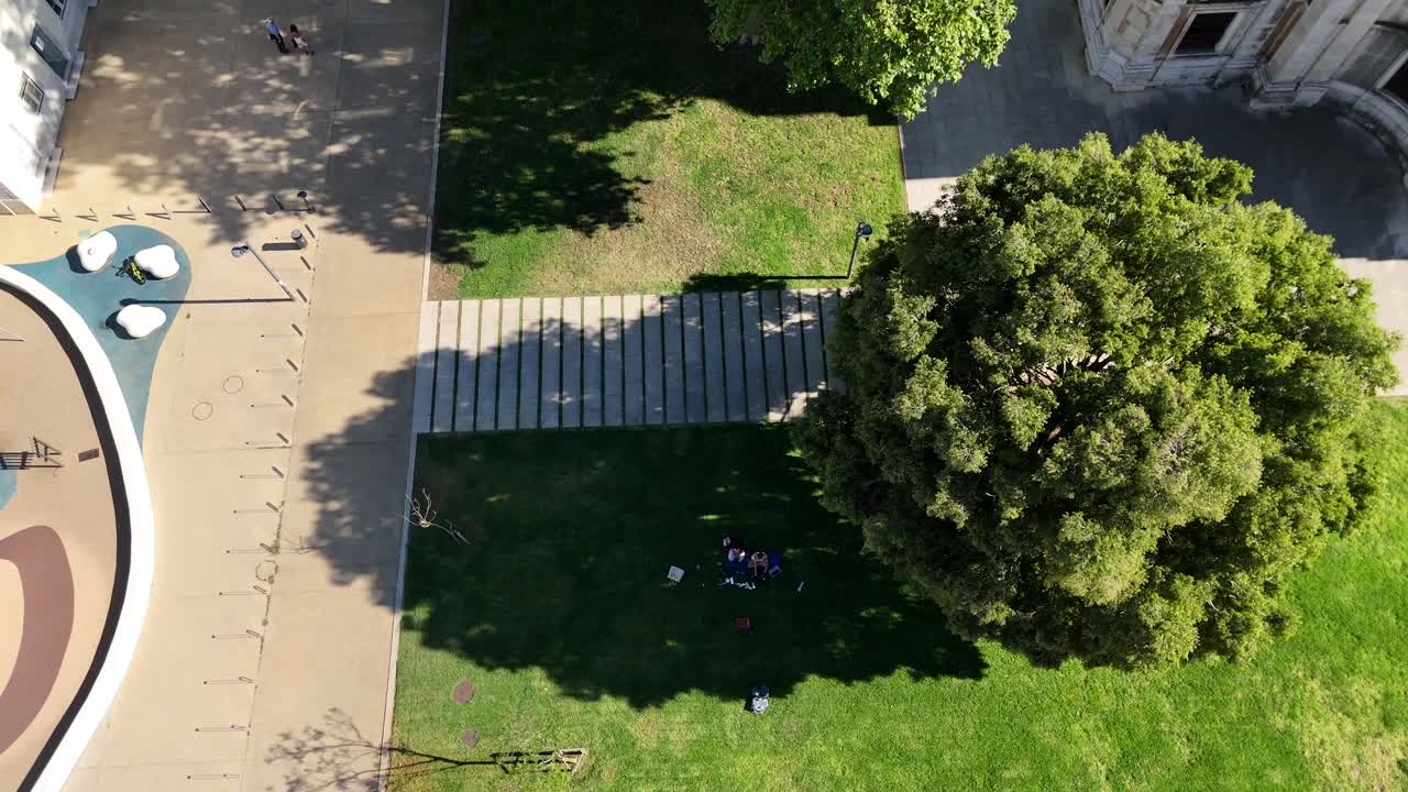 Overhead view of people relaxing and studying on the grass in a sunny urban park with pathways and trees, near buildings and a playground.