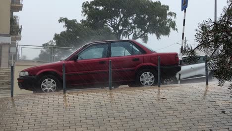 Extreme-Wind-And-Rain-hitting-red-car-parked-on-street,-Rainstrom-Concept