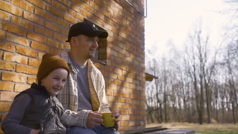 Caucasian-man-holding-a-cup-of-tea-sitting-with-her-son-outside-a-country-house