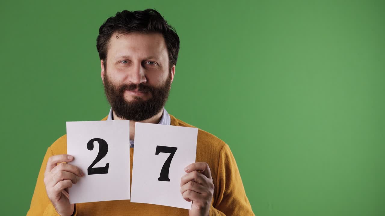 Man holding number 27 sign. Cheerful white bearded good-looking man in yellow sweater shows number twenty-seven on white sheet. Chrome key, green screen, copy space