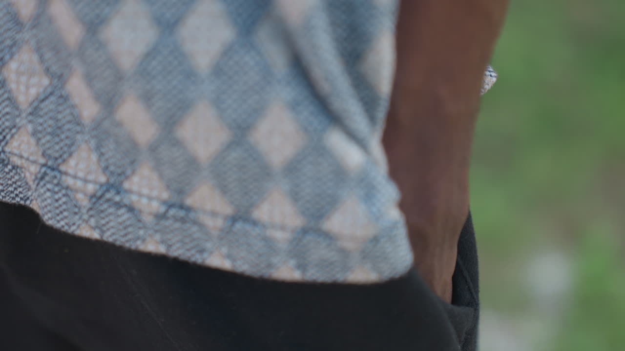 Black man adjusting hand in pocket, patterned top and black shorts. Tight outdoor closeup captures dark skin arm, intent posture, subtle movement suggesting pretraining focus, grass and sunlight