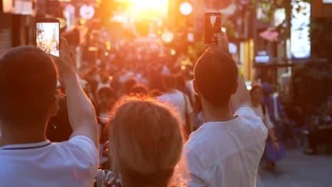 people taking pictures in a crowded city street at sunset