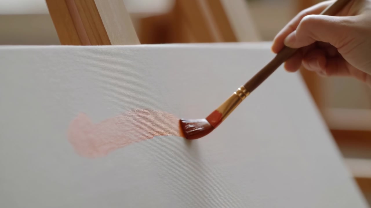 Pennello che dipinge rosa tenue su tela, primo piano di una mano che applica una sottile velatura acrilica con un pennello piatto su tela bianca