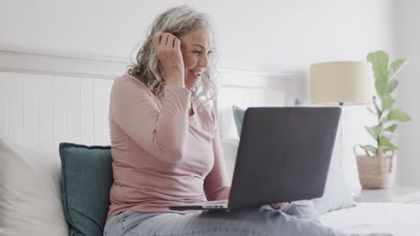 Glückliche-ältere-Kaukasische-Frau,-Die-Auf-Dem-Bett-Sitzt-Und-Laptop-Benutzt,-Zeitlupe