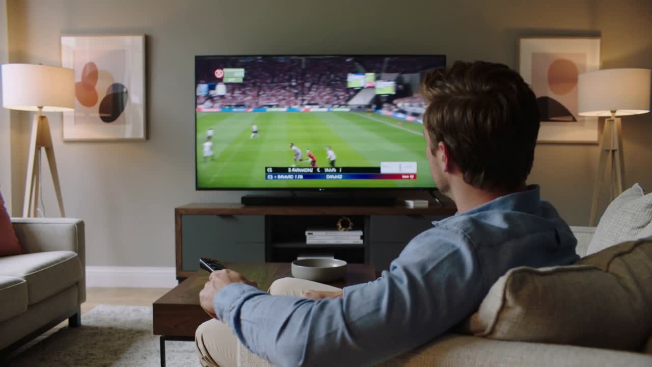 Uomo in camicia azzurra che reagisce alla partita in TV, regolando il telecomando sul divano del soggiorno, con spazio per il testo.