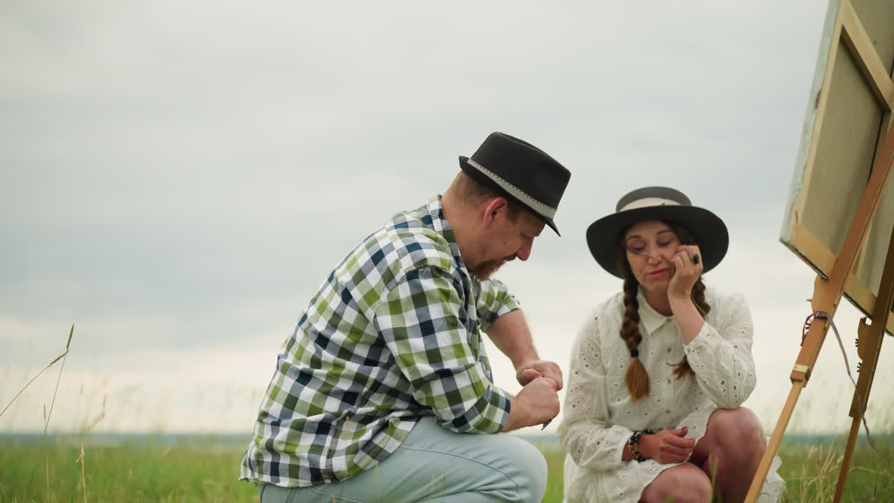 색 셔츠와 모자를 입은 남자가 페인트 튜브를 열기 위해 고군분투하는 동안 색 드레스와 모자 입은 여자가 보고 믿지 않고 머리를 흔들고 있습니다.